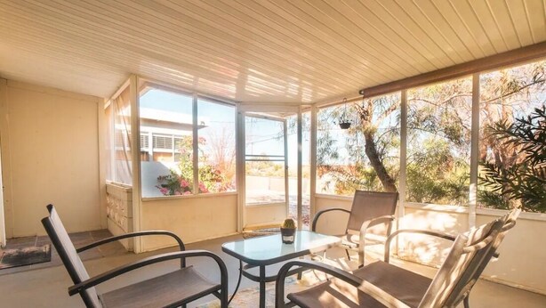 A sunlit patio with a table and chairs, large windows, and a view of the surrounding landscape. - Joshua Tree Desert Home (Joshua Tree)