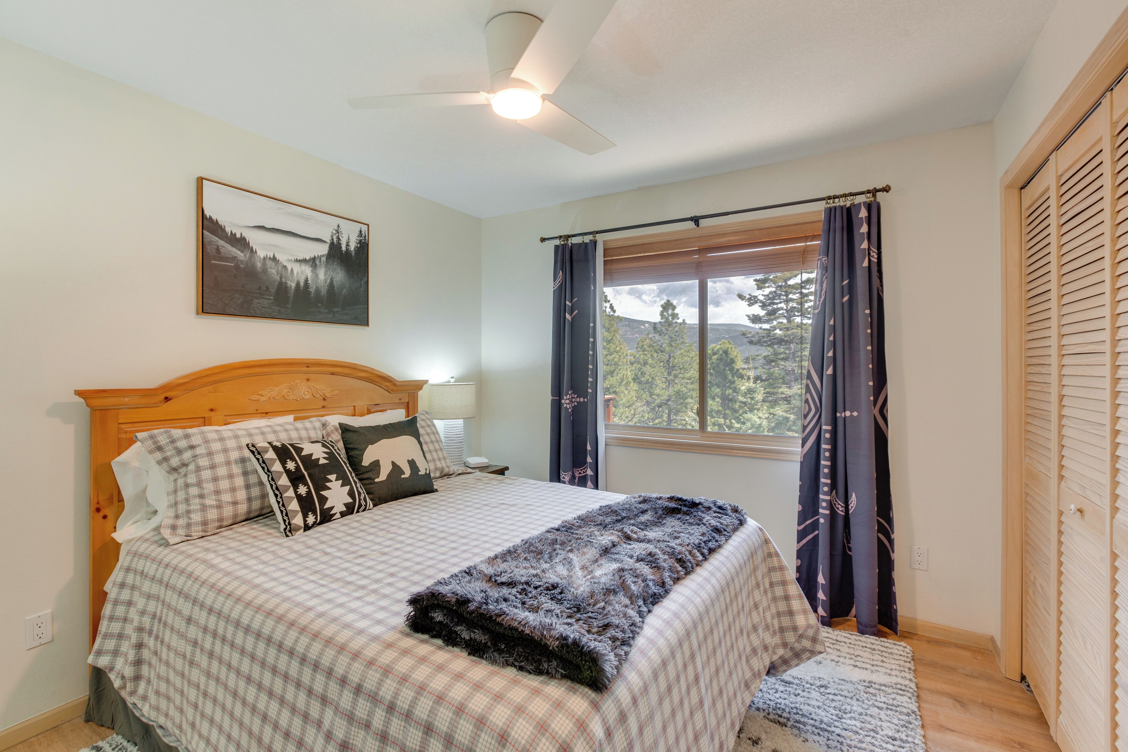 A bedroom with a bed, a ceiling fan, a window with curtains, and a door with louvered shutters.