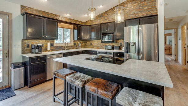 A modern kitchen with dark cabinets, a large island with stools, a stainless steel refrigerator, and a stone backsplash. - Angel Fire Rental Cabin w/ Hot Tub & Mountain View (Angel Fire)