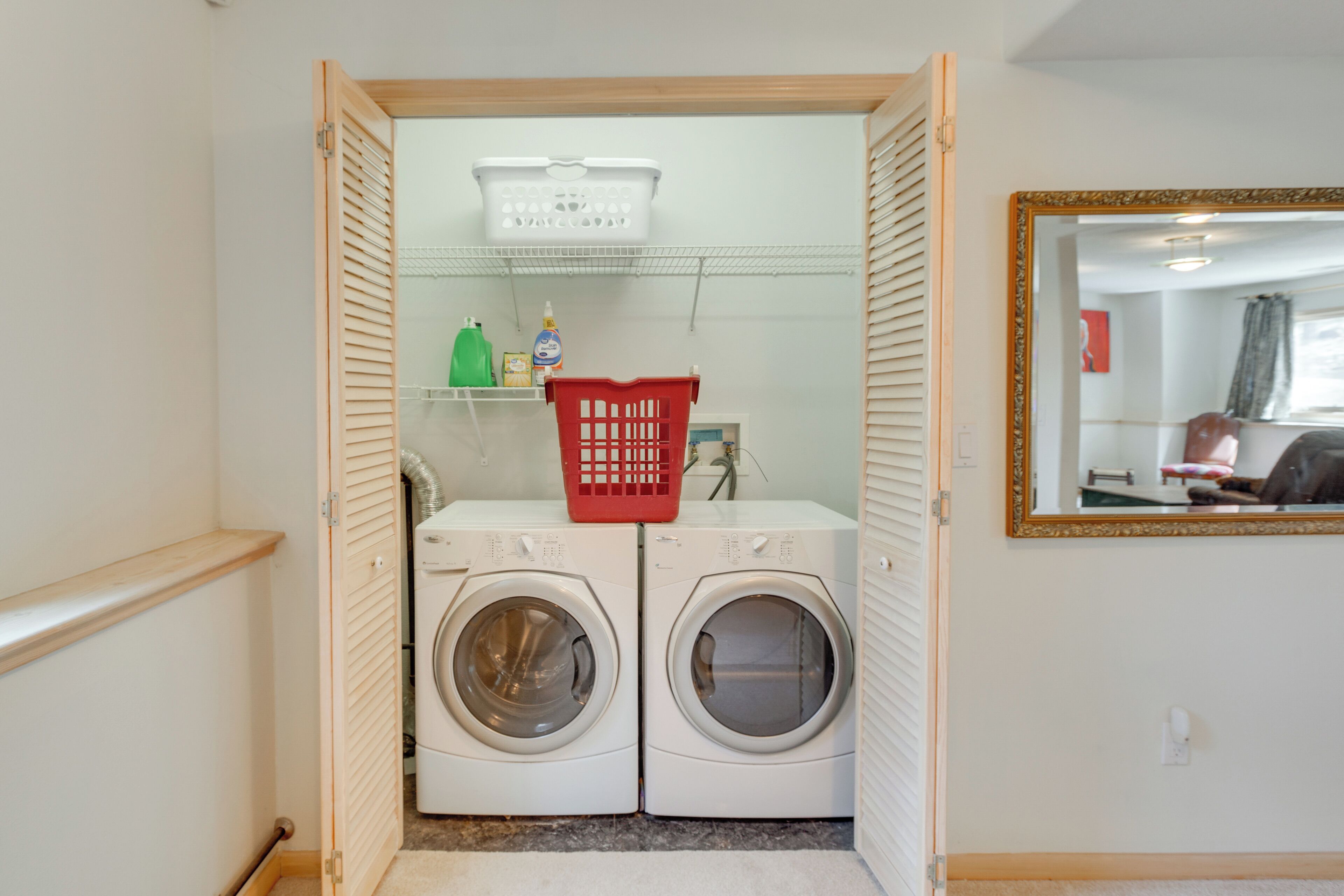 A laundry room with two front-loading washing machines, a red laundry basket, and shelves with cleaning supplies.