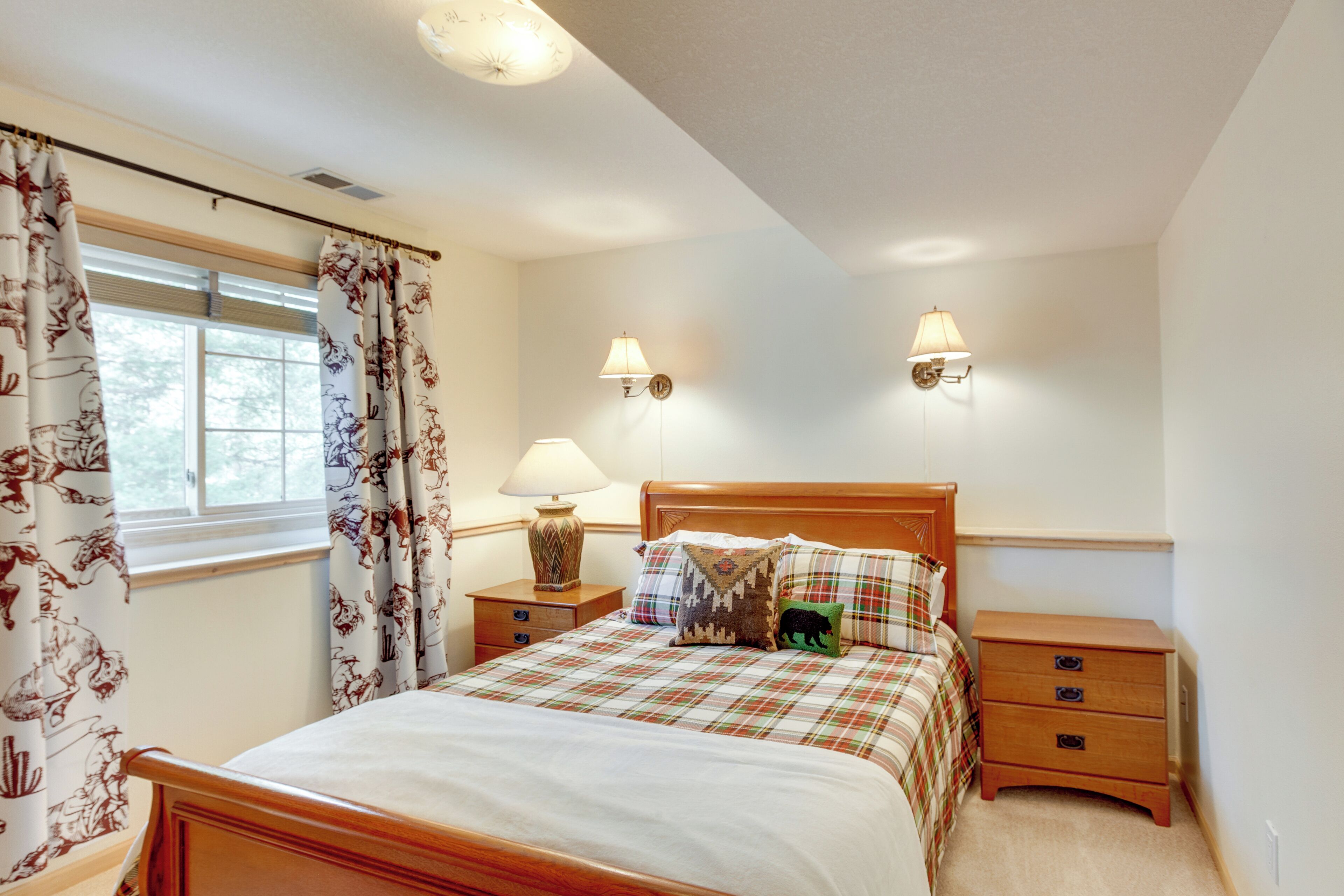 Een slaapkamer met een houten bed, twee nachtkastjes met lampen, een raam met bloemen gordijnen en een wandlamp.