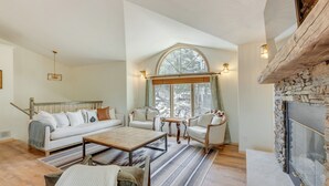 A cozy living room with a fireplace, a large window with curtains, and a wooden floor. - Angel Fire Rental Cabin w/ Hot Tub & Mountain View (Angel Fire)