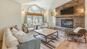 A living room with a stone fireplace, a large window, and a flat-screen TV mounted on the wall. - Angel Fire Rental Cabin w/ Hot Tub & Mountain View (Angel Fire)