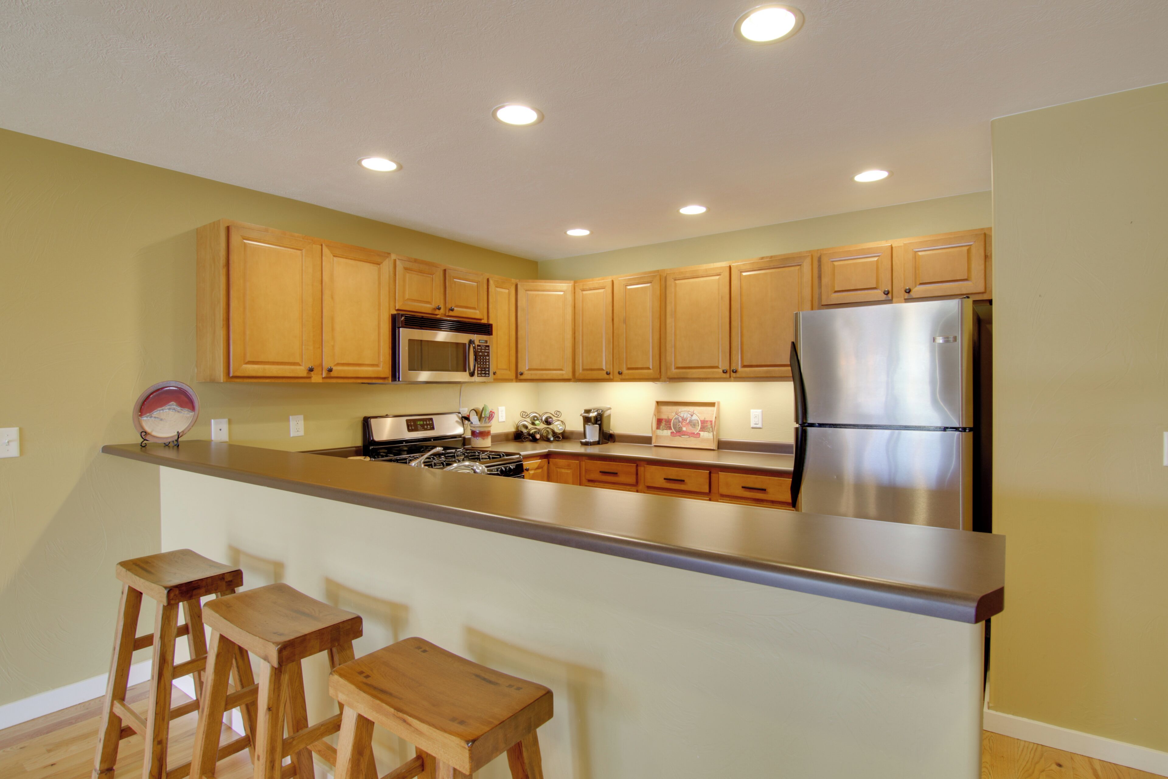 A kitchen with wooden cabinets, a stainless steel refrigerator, a microwave, and a stove. There are three wooden stools at the counter.