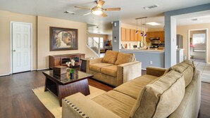 A spacious living room with a beige sofa, a glass coffee table, and a ceiling fan. A kitchen is visible in the background with wooden cabinets and a stainless steel refrigerator. - San Antonio Home w/ Private Hot Tub + Pool! (San Antonio)