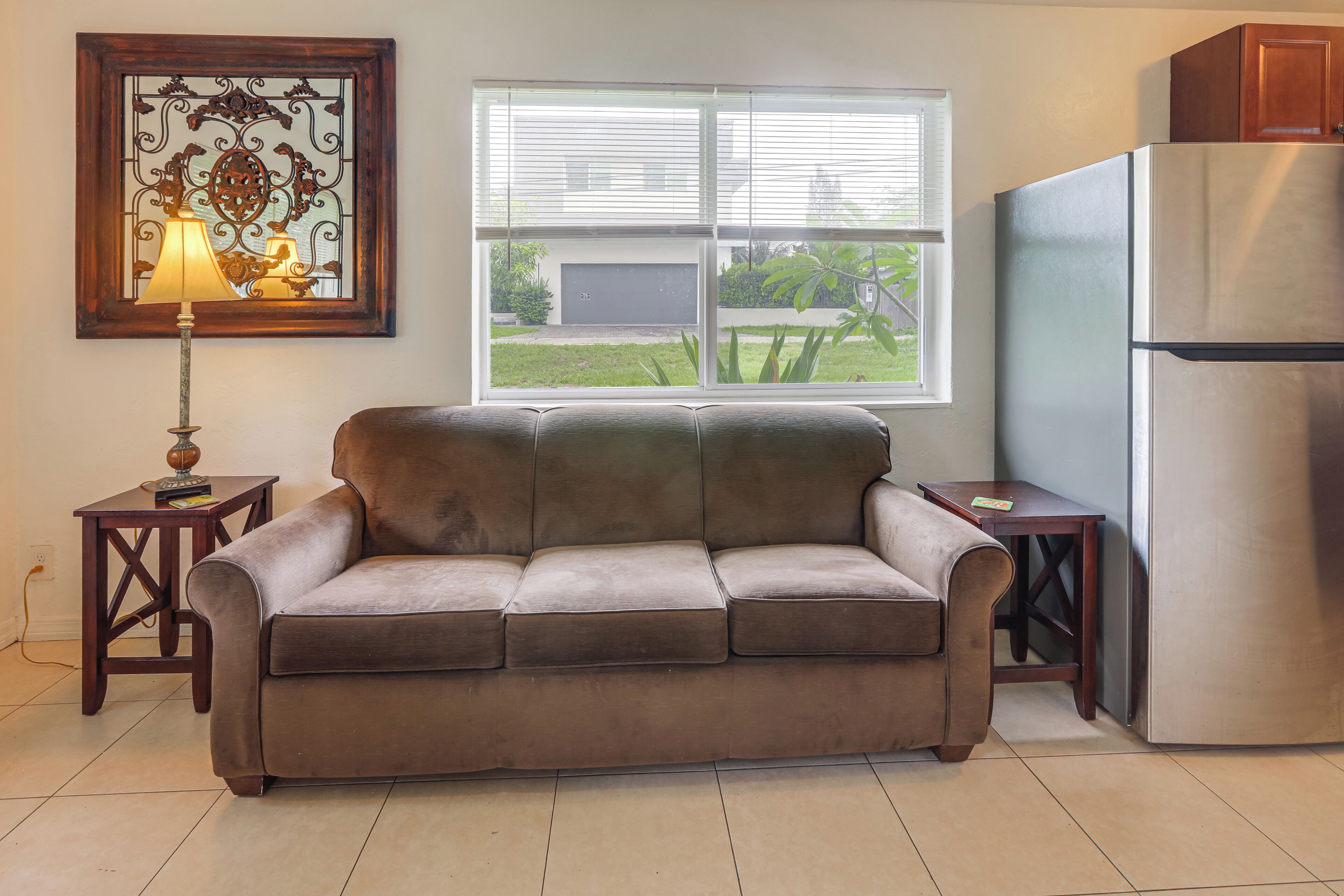 A living room with a sofa, a table, a lamp, a window with blinds, and a refrigerator.
