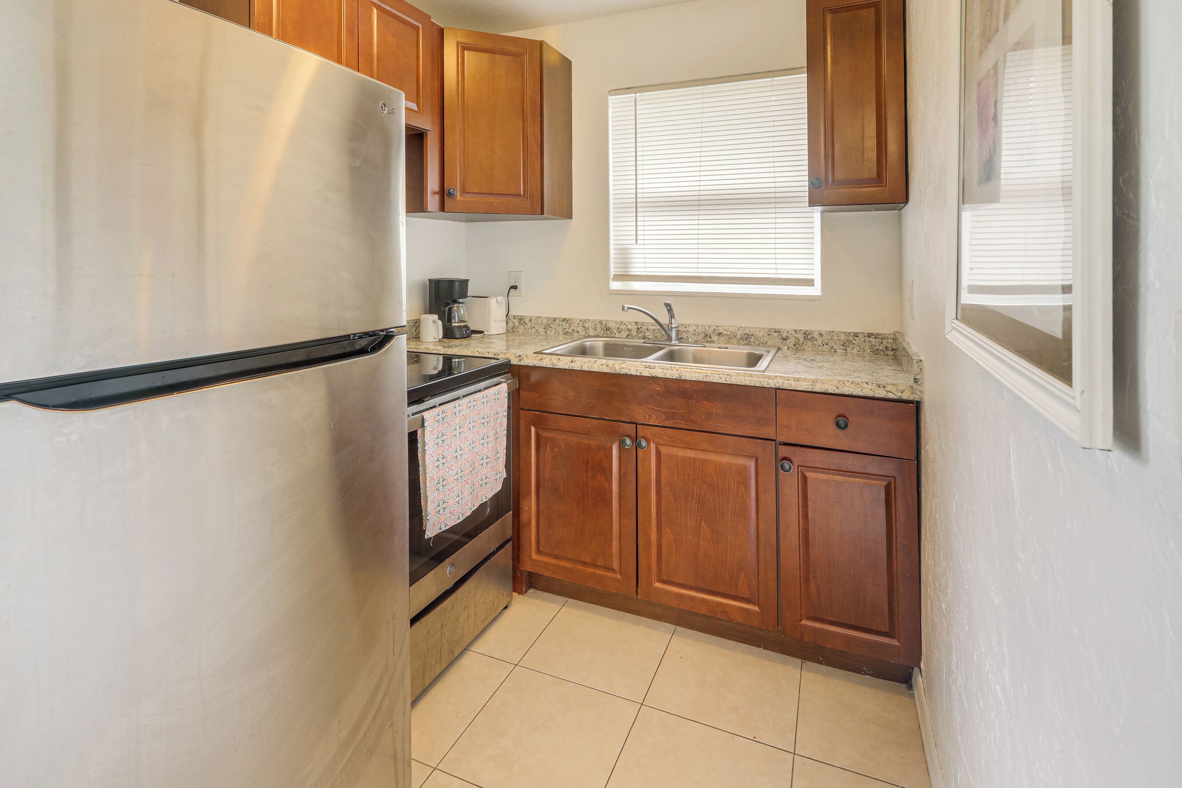 Una cocina compacta con armarios de madera, un refrigerador de acero inoxidable, un lavamanos doble y un ventanal con persianas.
