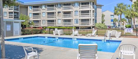 A swimming pool with white lounge chairs, a lifeguard station, and a multi-story building with balconies.
