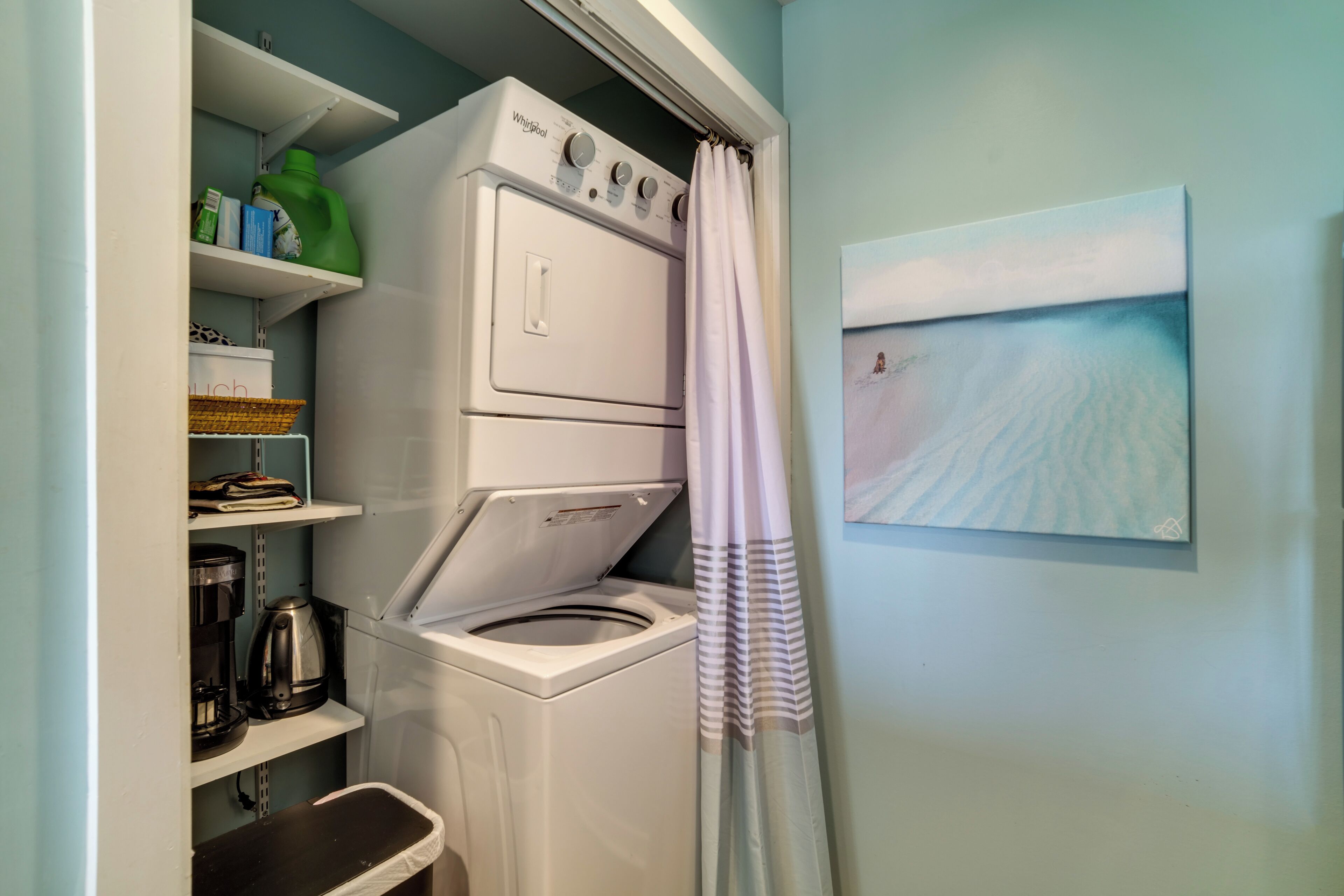 A compact laundry area with a front-loading washer and dryer, shelves with laundry supplies, a coffee maker, and a painting of a beach scene.