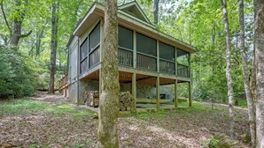 A two-story cabin with a balcony, surrounded by a forested area with a pile of firewood and a stone wall.