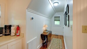 A narrow hallway with white paneled walls, a wooden floor, a small table with a lamp, a fire extinguisher, and a coffee maker.