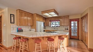 A kitchen with wooden cabinets, a central island with stools, a refrigerator, and a window with a view. - Near Bear Valley: Peaceful Arnold Home w/ Hot Tub (Arnold)