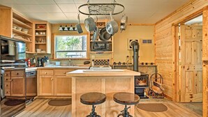 A cozy kitchen with wooden cabinets, a central island with stools, hanging pots, and a wood stove. - High Lakes Hideaway: La Pine Fishing Cabin! (La Pine)