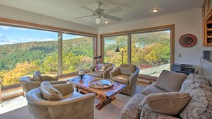 A spacious living room with large windows offering a view of a forested hillside. There are leather chairs, a wooden coffee table, and a sofa with patterned cushions.