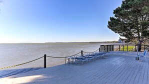 Een houten terras met stoelen gericht op een waterpartij, een touwbordeshek en een boom.