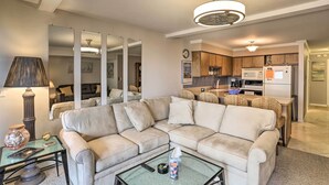 Een zithoek met een beige modulaire bank, een glazen salontafel en een lamp. Achtergrond: een keuken met houten kastjes en een koelkast.