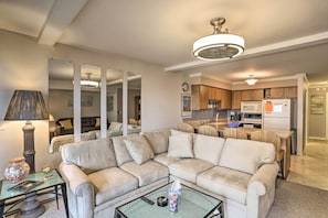 Una sala de estar con un sofá seccional color beige, una mesa de centro de vidrio y una lámpara. Se observa una cocina al fondo con gabinetes de madera y un refrigerador.