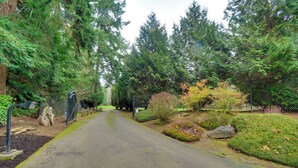 A paved pathway flanked by dense green foliage and a fence. - Central 4-acre Cottage w/ Deck: Walk to Bay! (Bainbridge Island)