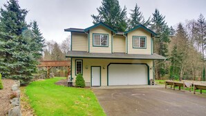 A two-story house with a garage, surrounded by a well-maintained lawn and trees. - Central 4-acre Cottage w/ Deck: Walk to Bay! (Bainbridge Island)