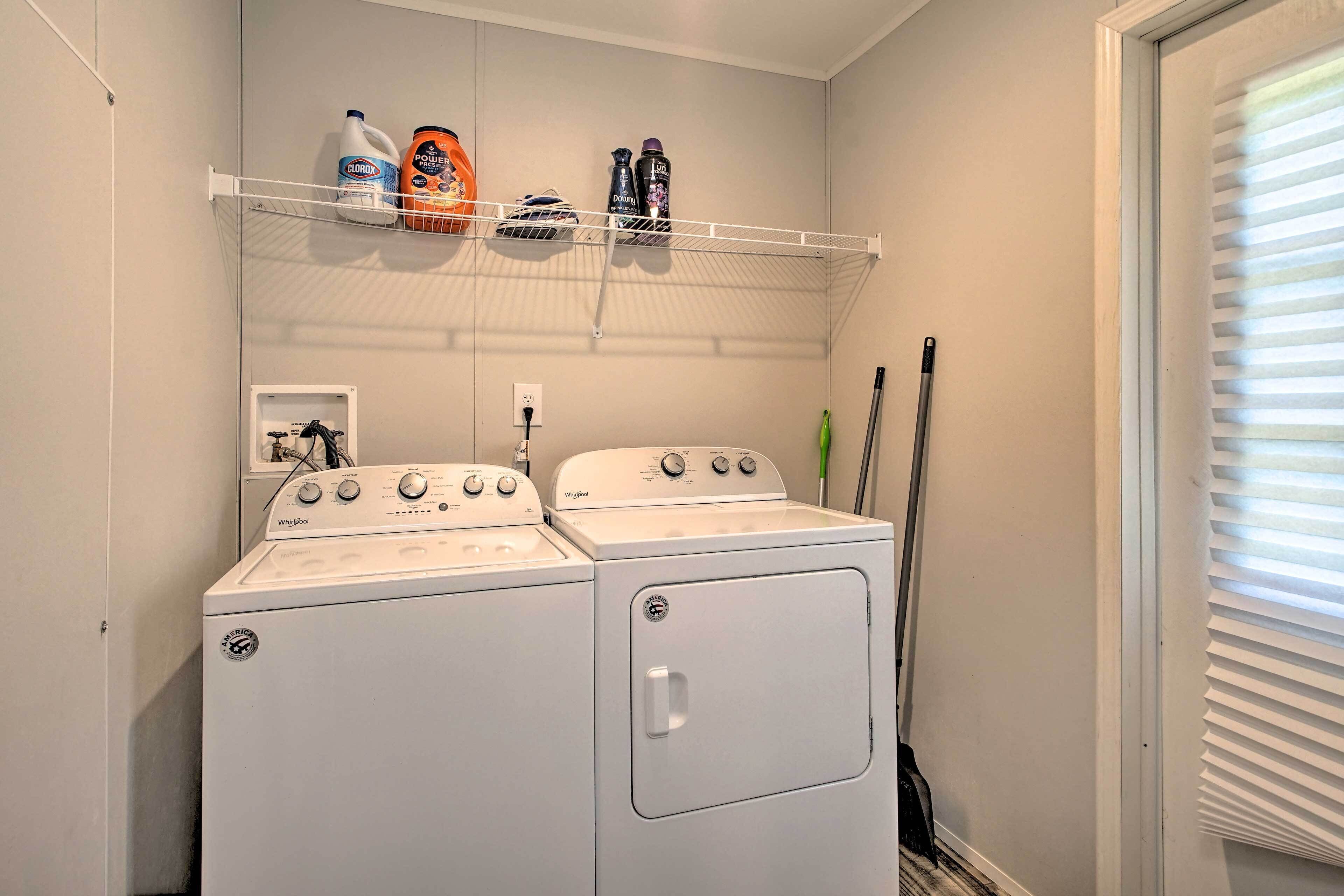 Une buanderie équipée de deux lave-linge et deux sèche-linge Whirlpool, d’une étagère contenant des produits de nettoyage et d’une fenêtre avec des stores.