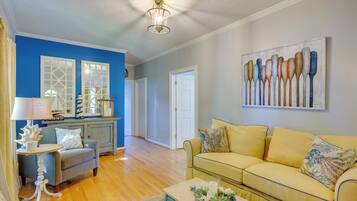 A living room with a blue accent wall, a yellow sofa, a white table with a lamp, and a painting of spoons.