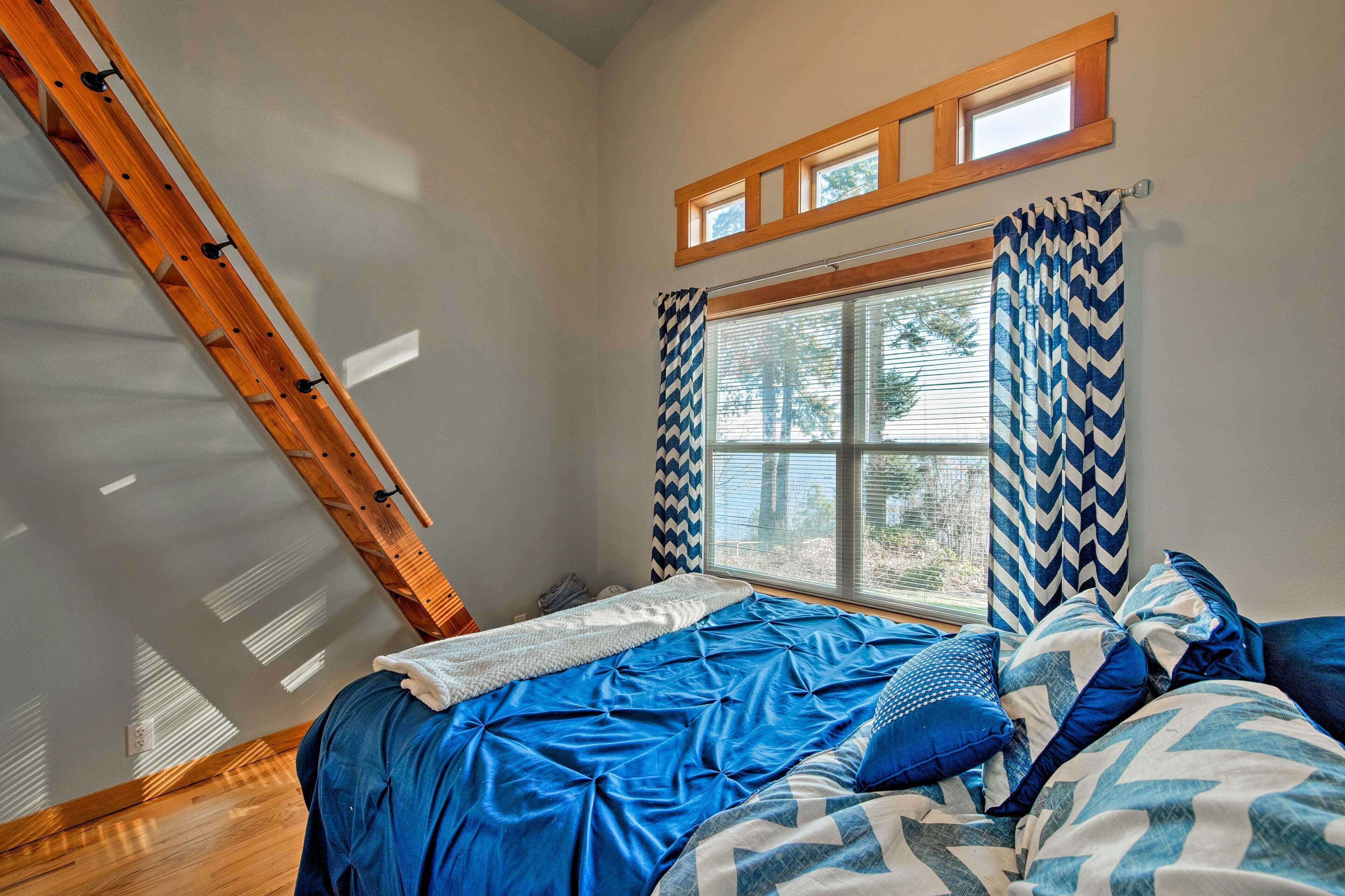 Een slaapkamer met een schuine plafond, een bed met blauw beddengoed, een raam met ruitpatroon gordijnen, en een houten ladder.