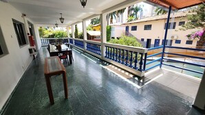 Un espacio al aire libre techado con una mesa y sillas, barandilla azul y vistas a edificios y follaje.