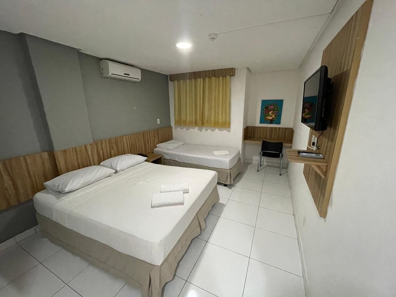 Quarto de hotel com duas camas, televisão, pequena escrivaninha e janela com cortinas.