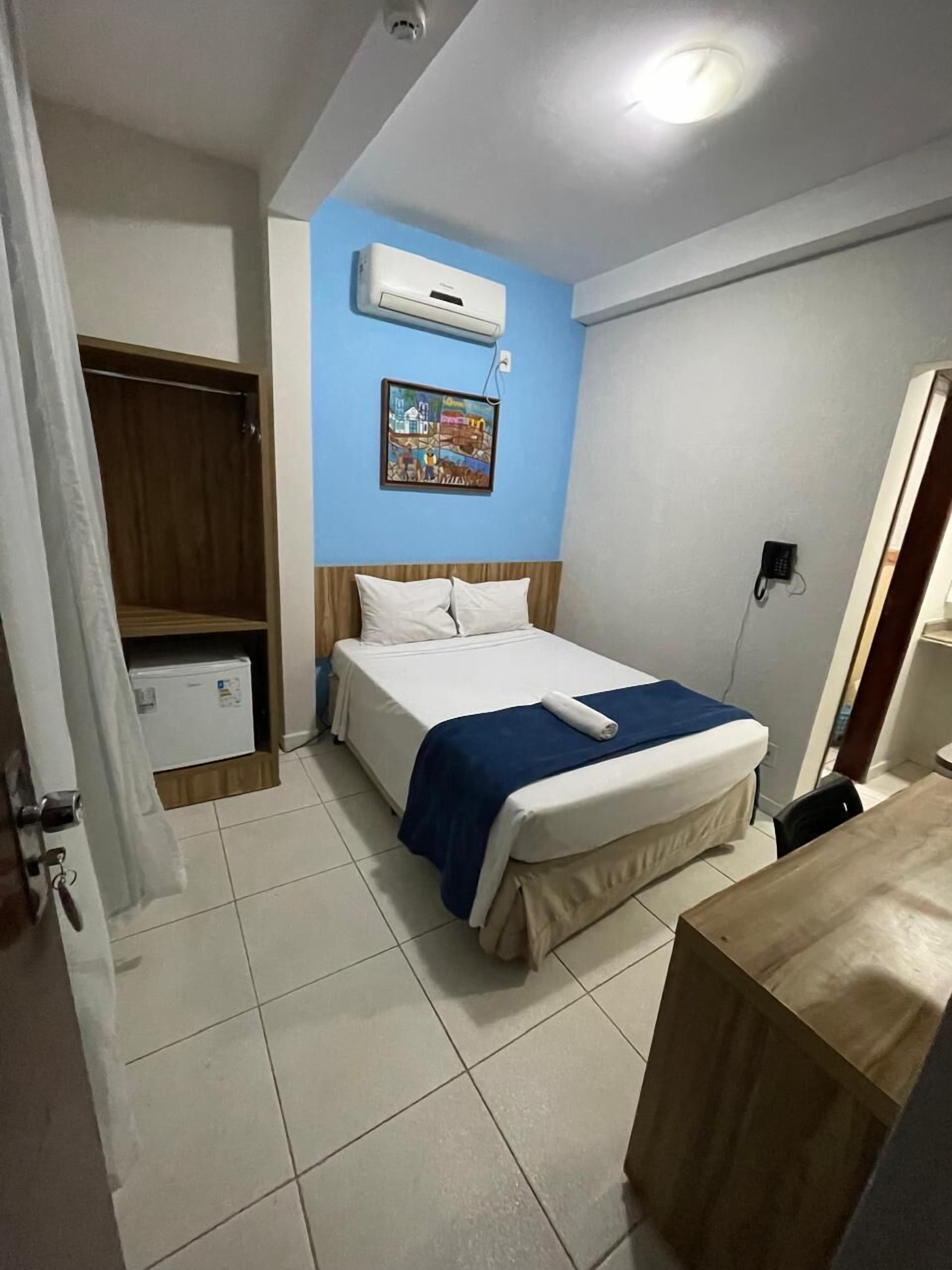 A hotel room with a bed, a wardrobe, a small table, a wall-mounted air conditioner, and a painting.