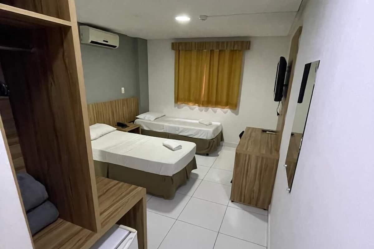 Une chambre d’hôtel compacte comprenant deux lits simples, un petit réfrigérateur, une armoire en bois et une fenêtre avec des rideaux.