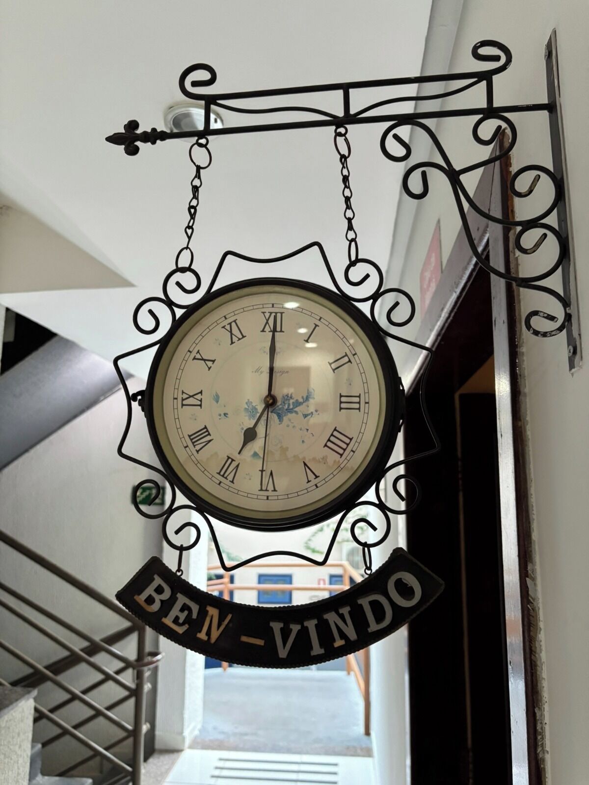 A wall-mounted clock with Roman numerals and a decorative frame, hanging above a staircase with a sign that reads "BEN-VINDO".