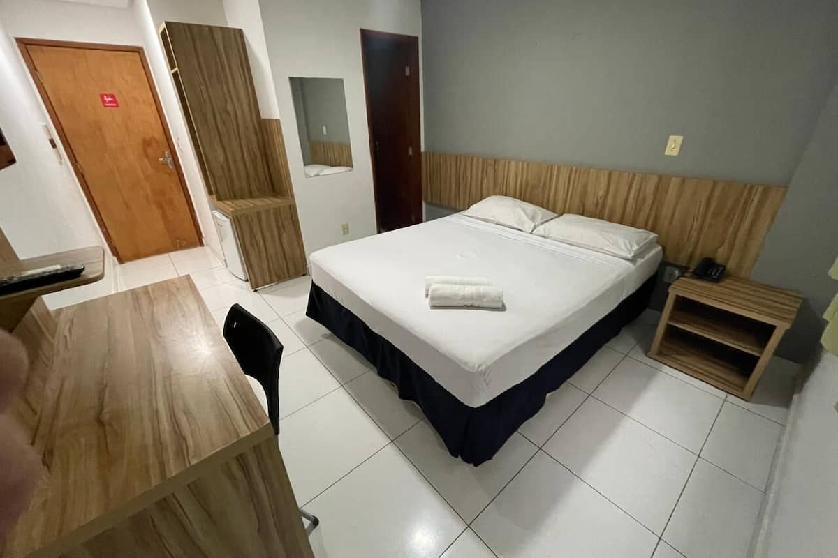 Une chambre d’hôtel comprenant un lit, un bureau, une chaise, une armoire et une petite table de chevet en bois.