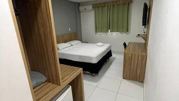 Habitación de hotel pequeña con cama, escritorio, refrigerador, televisor y ventana con cortinas.