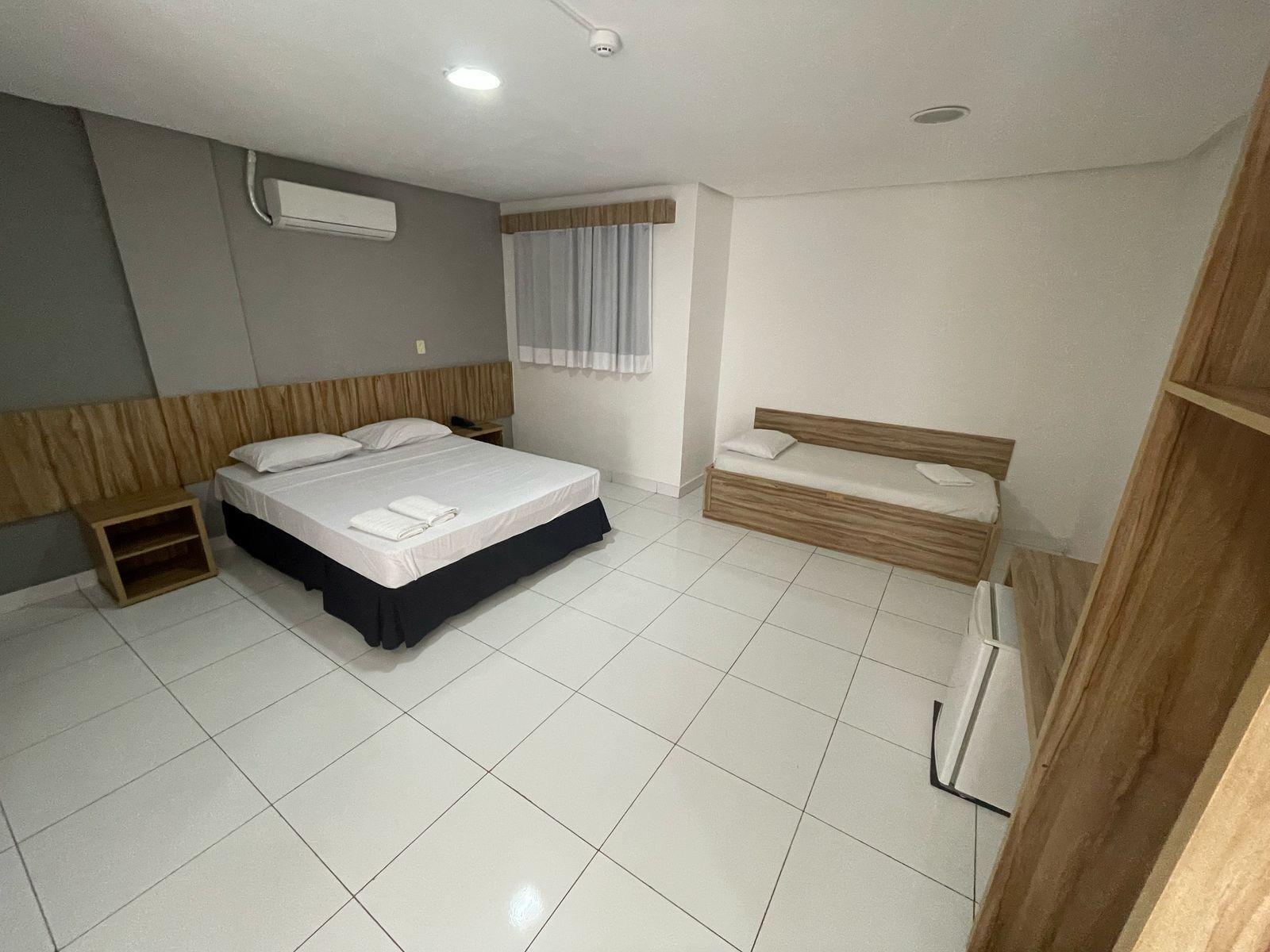 Quarto de hotel com uma cama grande, um sofá menor, uma cabeceira de madeira, uma mesinha de cabeceira e uma janela com cortinas.
