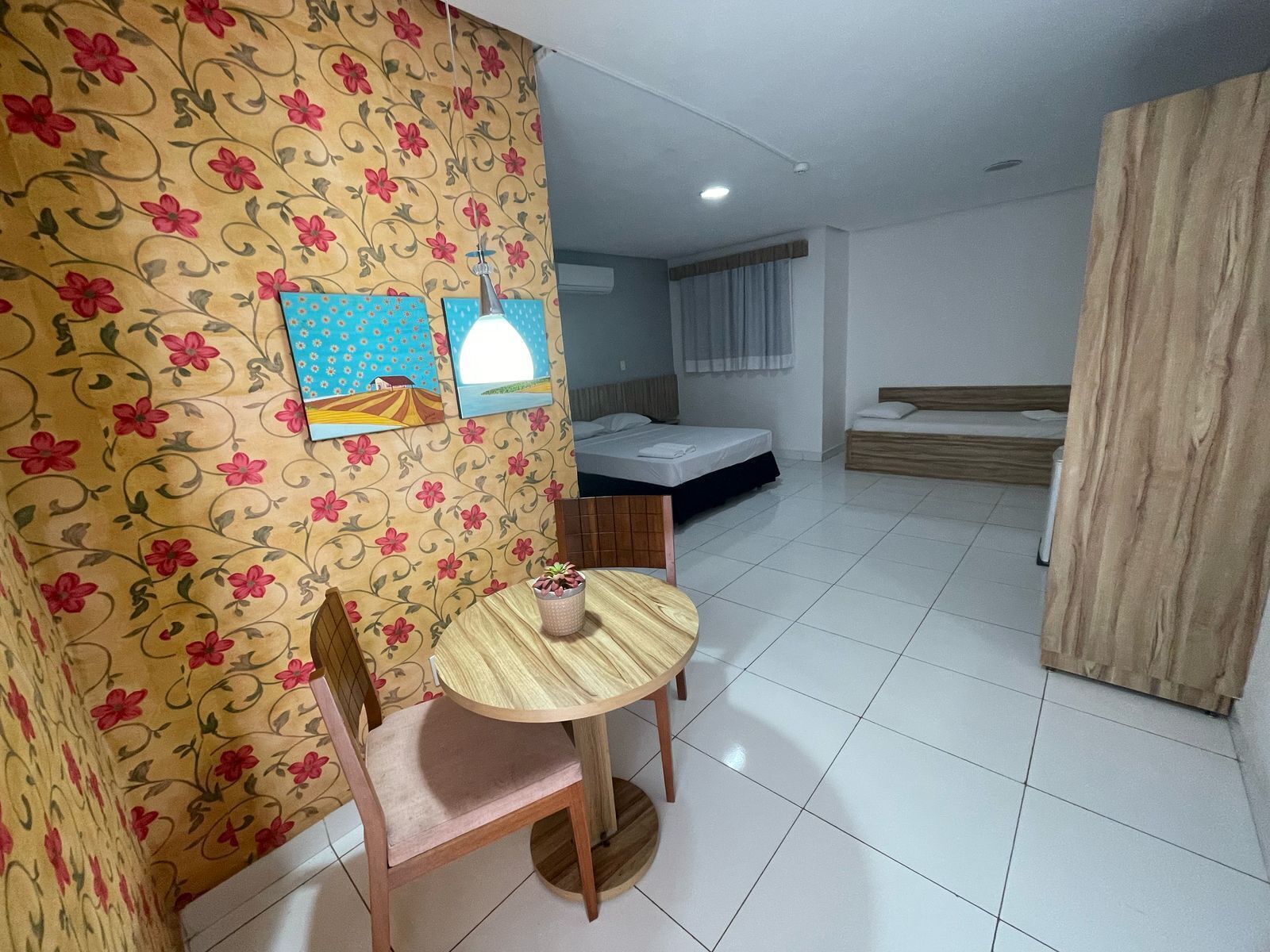 A compact hotel room with a bed, a small table with a chair, a wall with floral wallpaper, and a window with curtains.