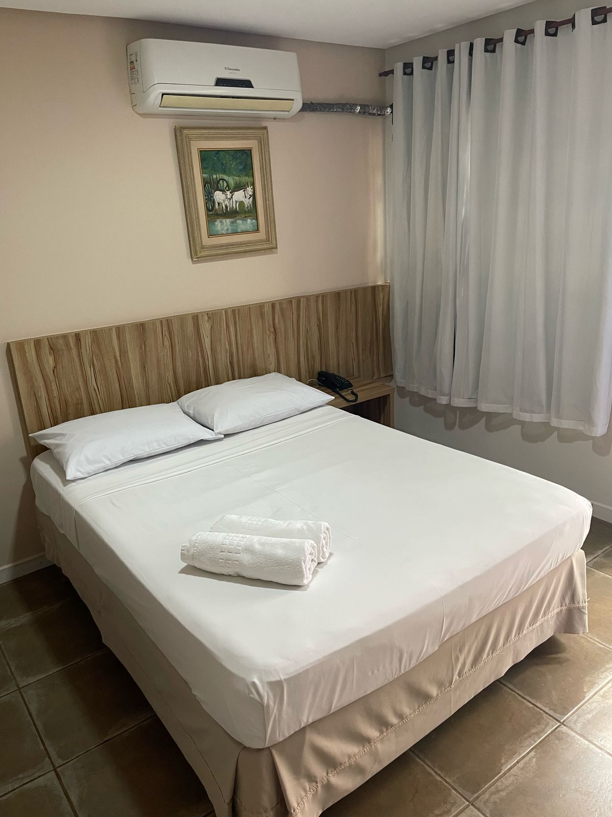 Habitación de hotel con cama, teléfono, aire acondicionado, un cuadro y ventana con cortinas.