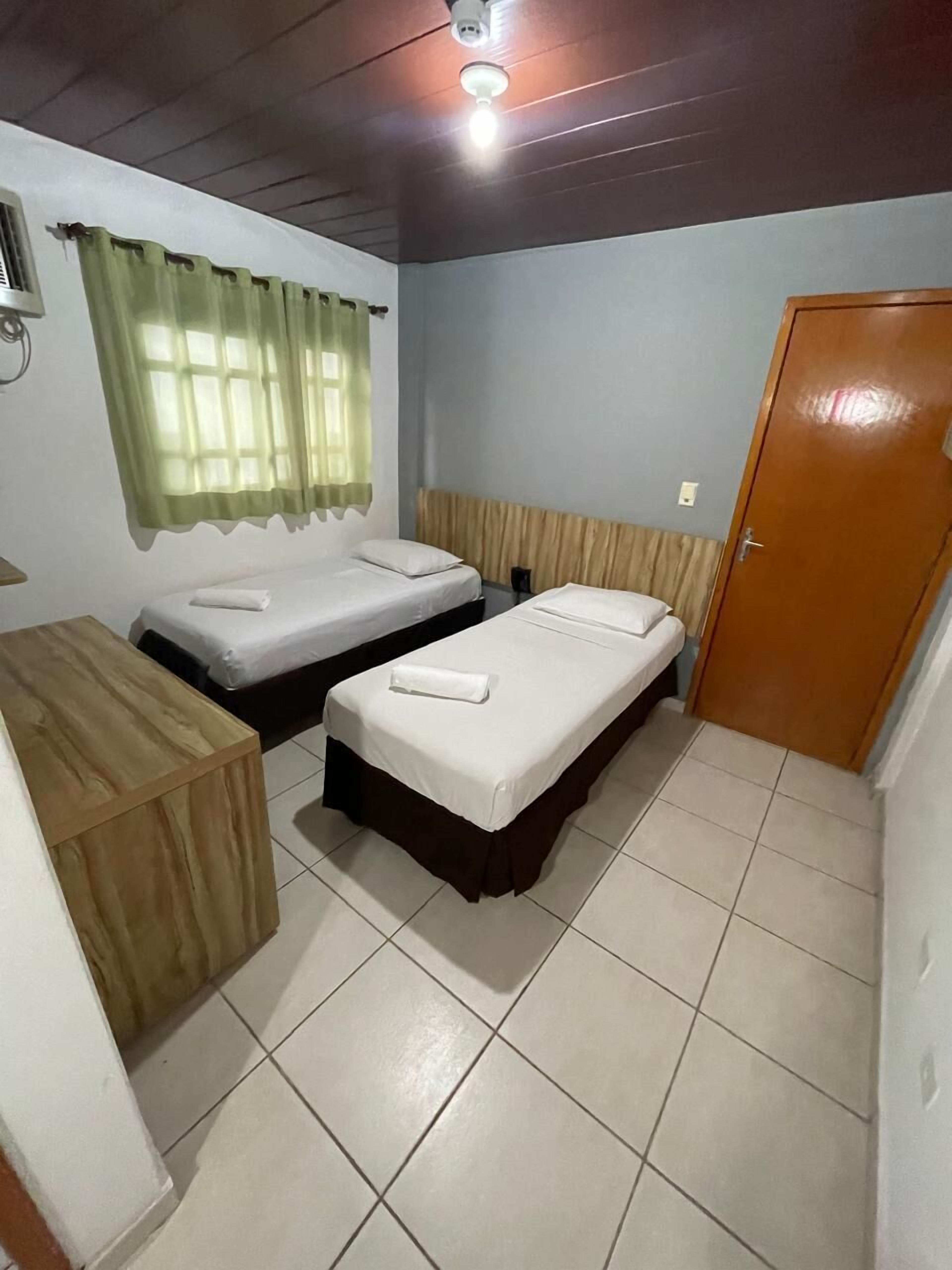 Quarto com duas camas de solteiro, uma escrivaninha de madeira, uma porta e uma janela com cortinas.