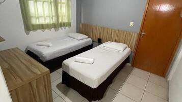 Quarto com duas camas de solteiro, uma escrivaninha de madeira, uma porta e uma janela com cortinas.