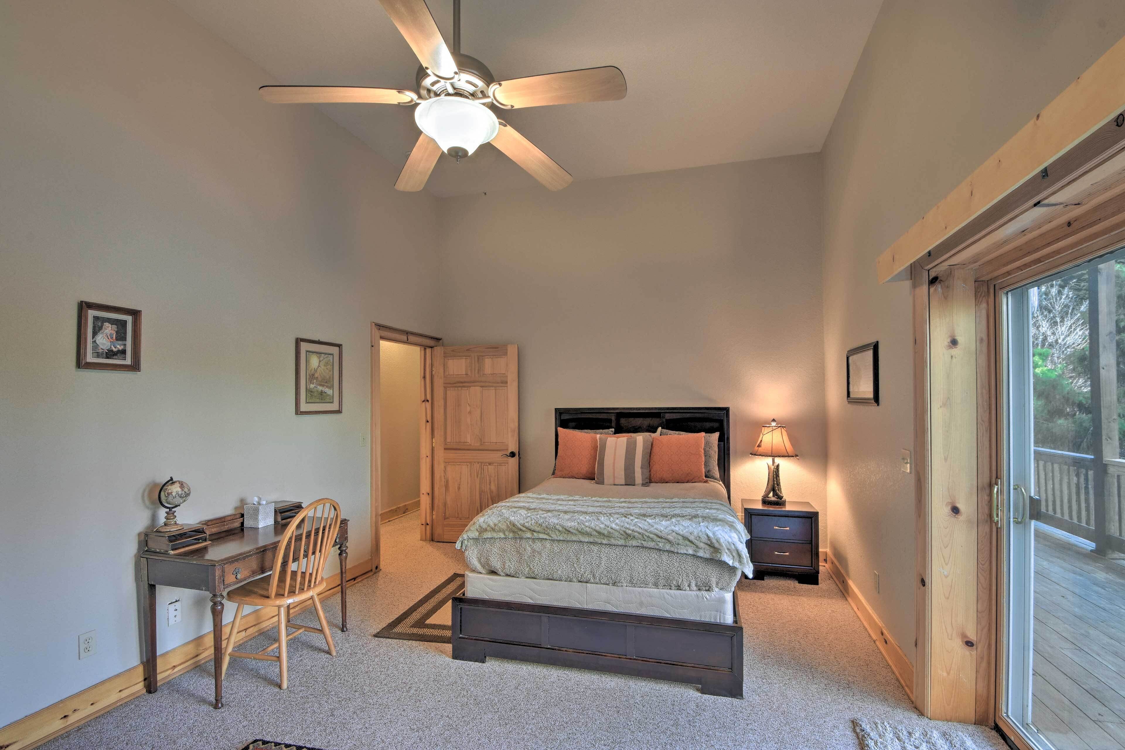 A bedroom with a bed, desk, chair, ceiling fan, and a view through a window.