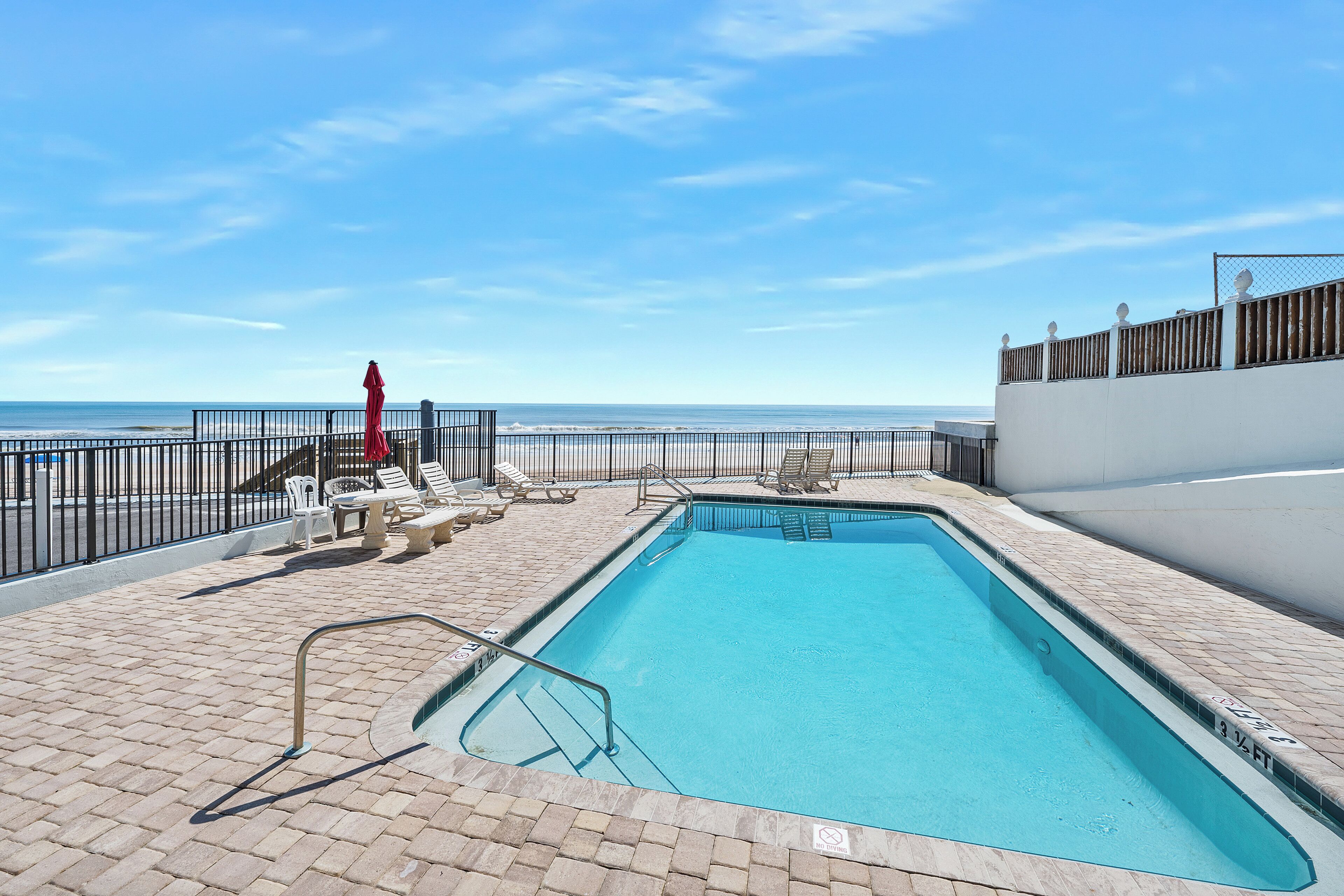 A rectangular pool with a metal ladder, surrounded by a brick patio, white lounge chairs, and a red umbrella.