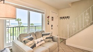 Uma sala de estar com sofá bege, uma janela grande com vista para o oceano e escada com corrimãos brancos.