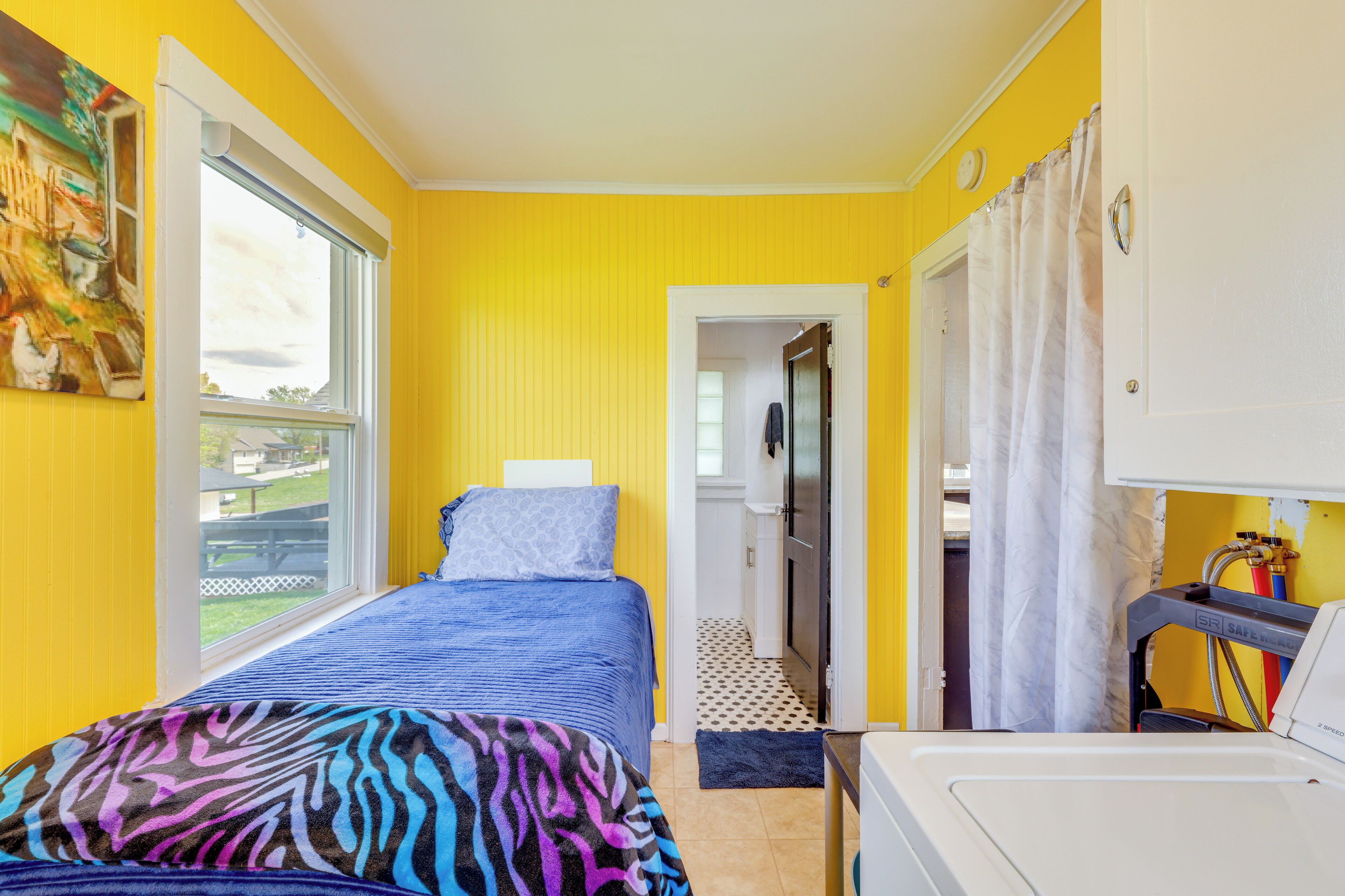 A room with yellow walls, a bed with a blue and zebra-patterned blanket, a white washing machine, and a view of a garden through a window.