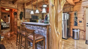 L’intérieur d’un chalet rustique est agrémenté d’une cheminée en pierre, de tabourets de bar en bois, d’un coin cuisine avec un réfrigérateur et d’un mur en pierre.