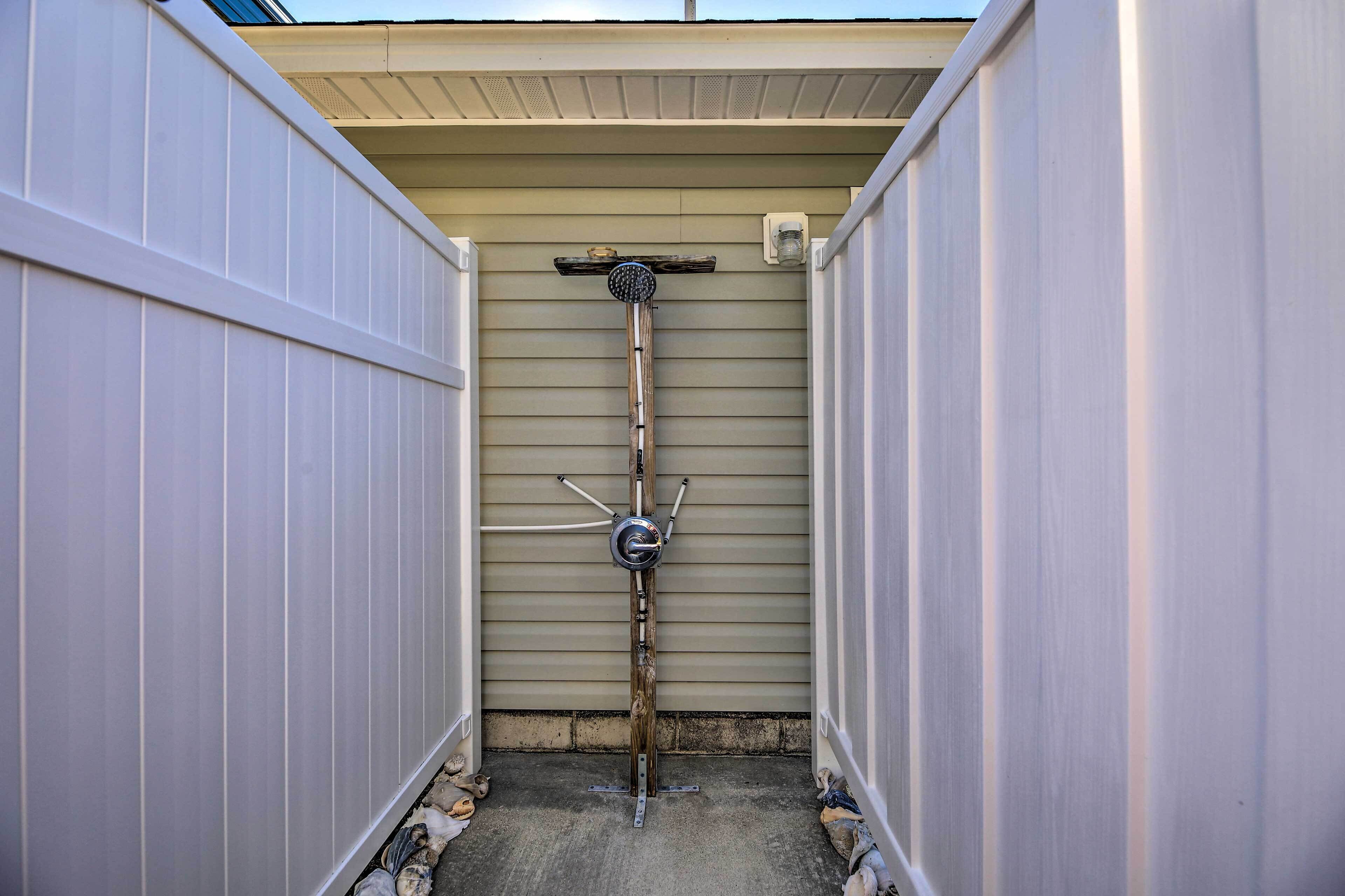 A narrow outdoor shower area with a metal showerhead mounted on a wooden post, surrounded by white siding walls.