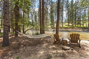 Un'amaca tra gli alberi, una sedia di legno e un tavolo da picnic in una zona boschiva con un fiume.