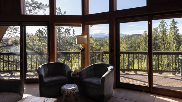 A spacious balcony with large windows offering a view of a forest and mountains. There are two armchairs and a small round table with a lamp.