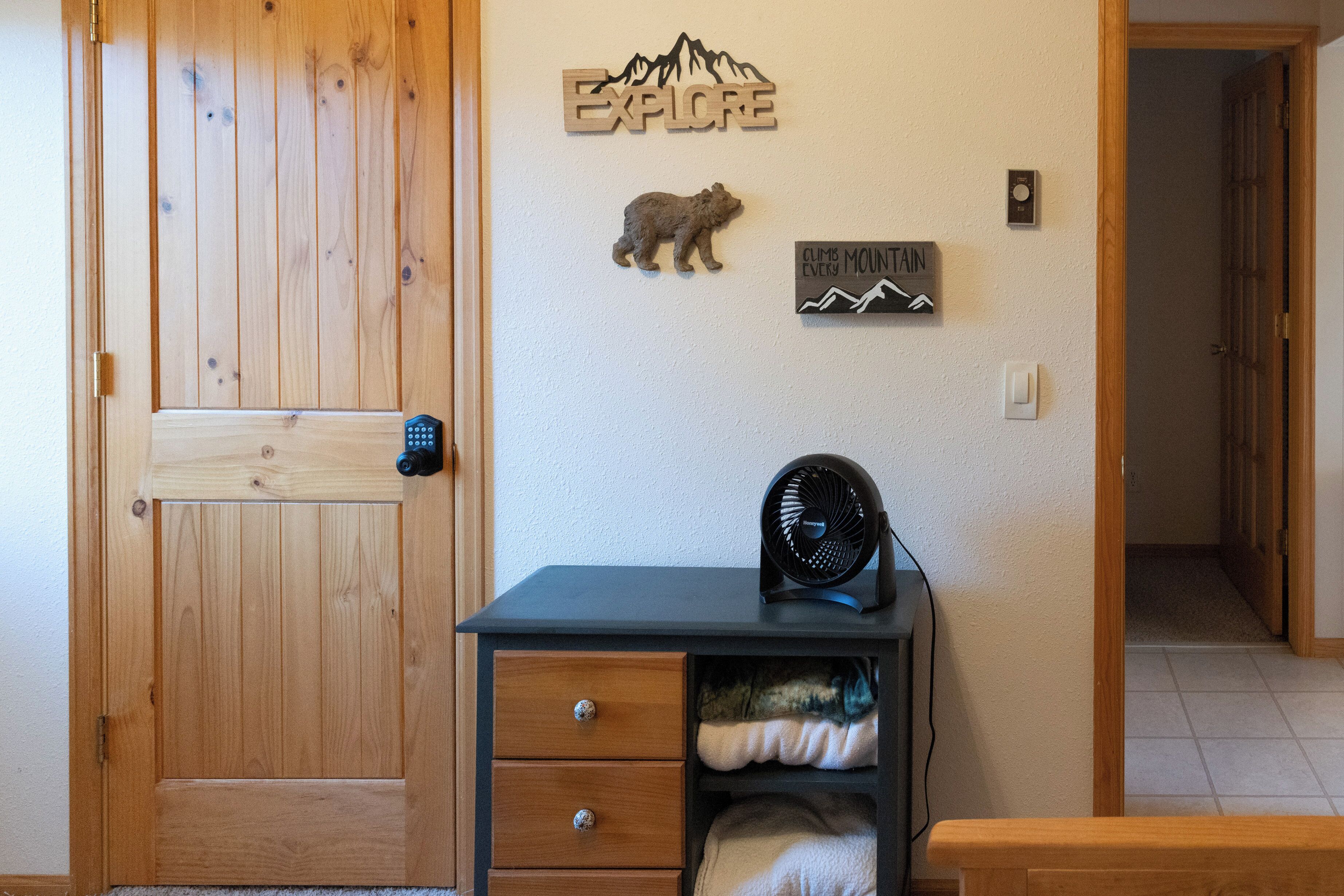 Een kamer met een houten deur, een klein tafeltje met een ventilator, en decoraties aan de muur, waaronder een beer en een bord waarop 'EXPLORE' staat.