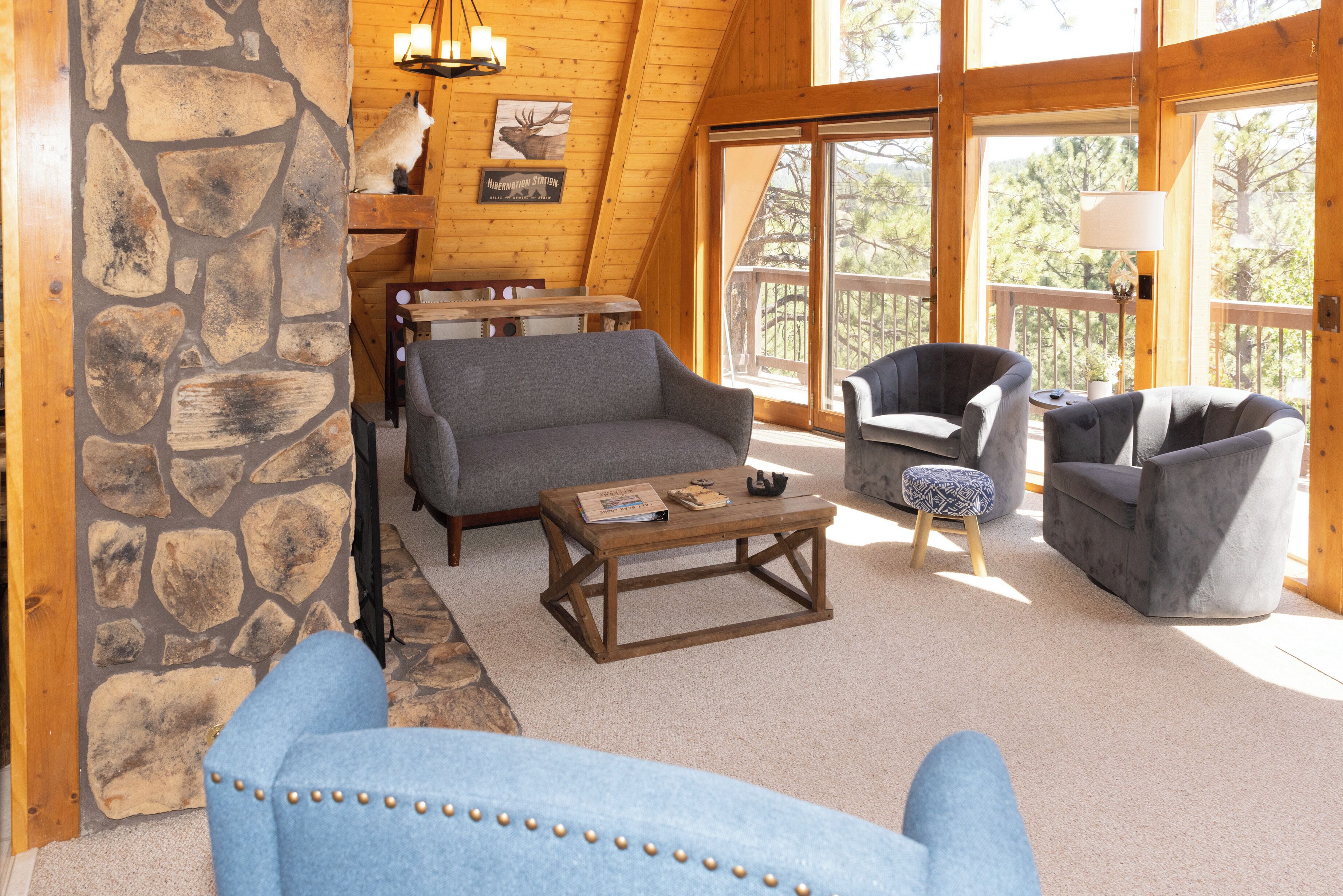 A cozy cabin interior with a stone wall, wooden ceiling, and large windows. There are two sofas, a coffee table, and a small table with a lamp.