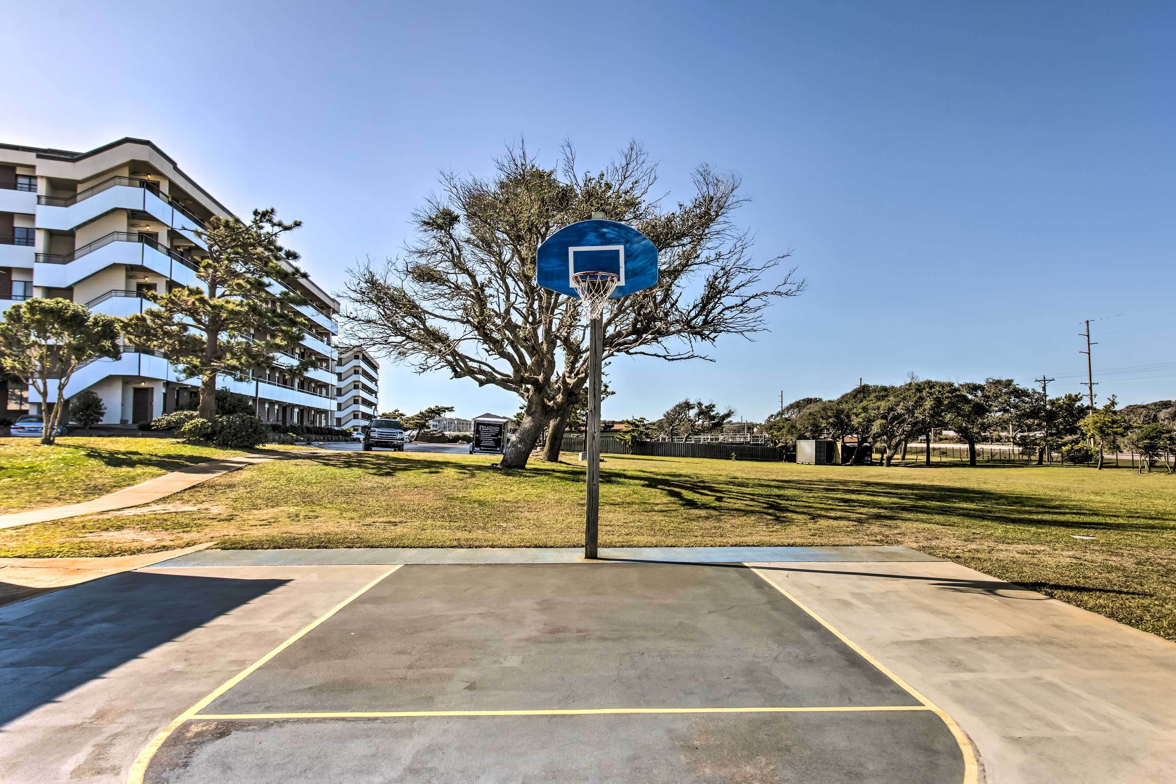 Una cancha de baloncesto al aire libre con un aro azul, rodeada de césped y un edificio de varios pisos al fondo.