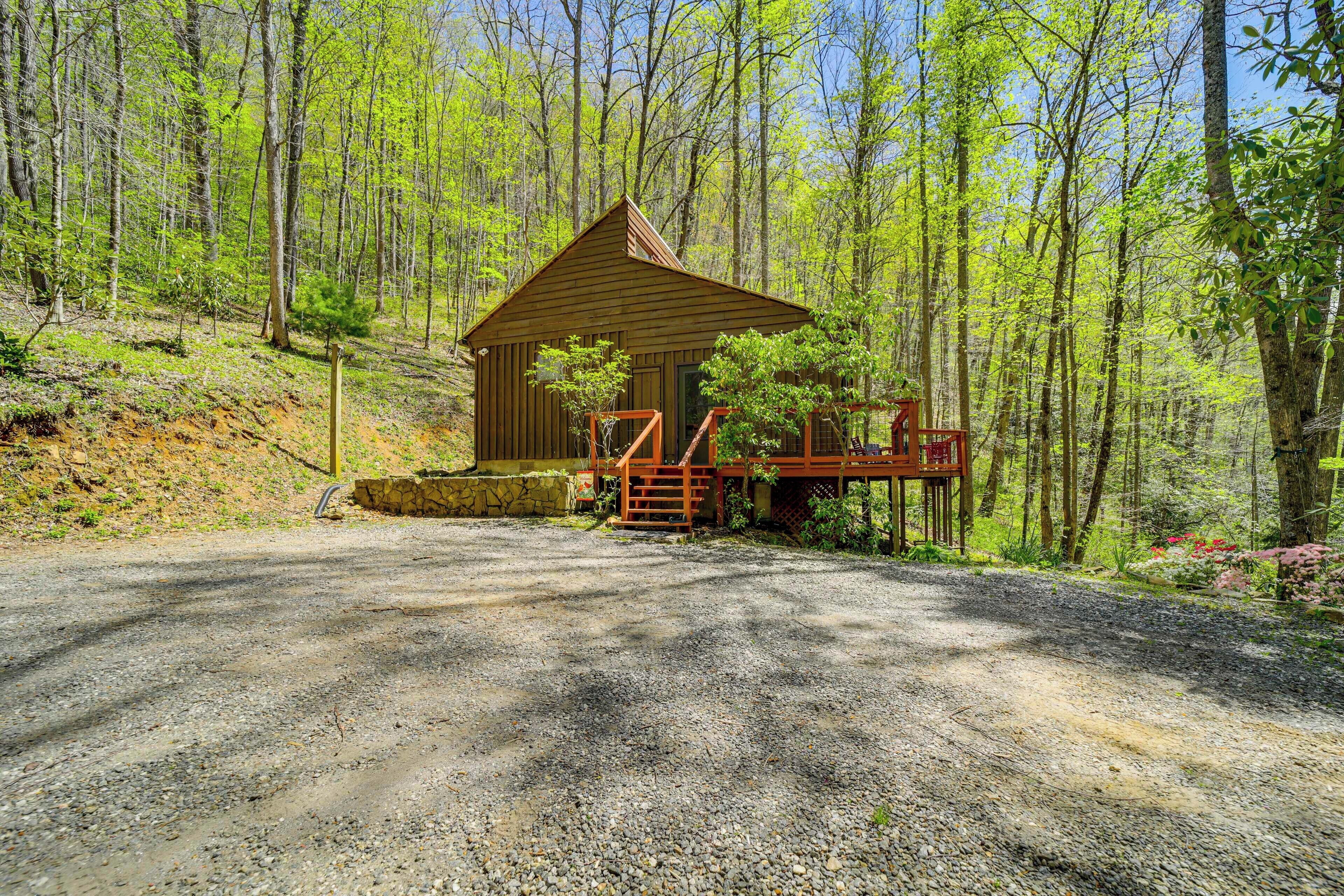 A wooden cabin with a porch and a gravel driveway in a forested area.